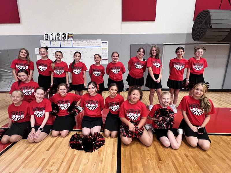 a group of middle school girls in tshirts and swirly skirts, sitting in a posed group with poms and their coach